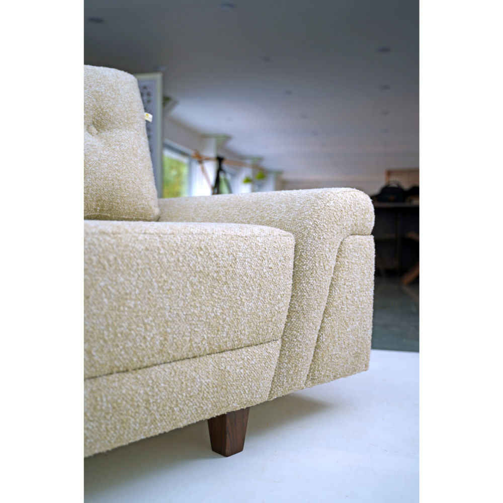 A light beige Meta Boucle sofa from Tooba Furniture Factory, featuring three back cushions, is displayed against a white background. This three-seater sofa boasts a modern design with plush upholstery and slightly angled armrests. It rests on four short, dark wooden legs.