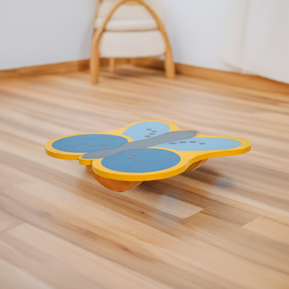 A brightly colored Butterfly balancing board from the Tooba Furniture Factory rests on a polished hardwood floor. The butterfly design, painted in blue and yellow, creates an engaging children's toy that encourages physical activity. In the background, there is a white wall and blurred furniture, including a white cushioned chair.