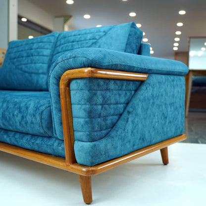 The Quantum Sofa by Tooba Furniture Factory is a modern teal upholstered piece that boasts a wooden frame and legs. It features simple, clean lines, cushioned armrests, and an angled backrest with a quilted design. Wooden accents enhance its stylish and contemporary appeal.